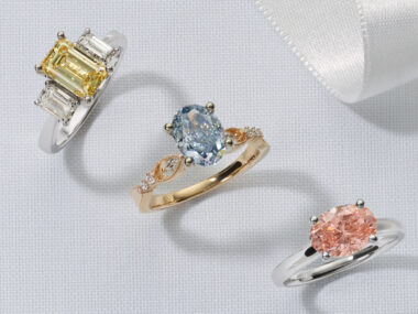 3 rings featuring Fancy Color lab-grown diamonds. An emerald cut fancy yellow lab-grown diamond in a white gold three stone ring with diamonds, an oval cut fancy blue lab-grown diamond set in a gold ring, and an oval shape fancy pink lab-grown diamond set in a white gold ring