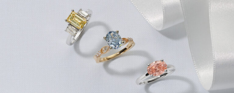 3 rings featuring Fancy Color lab-grown diamonds. An emerald cut fancy yellow lab-grown diamond in a white gold three stone ring with diamonds, an oval cut fancy blue lab-grown diamond set in a gold ring, and an oval shape fancy pink lab-grown diamond set in a white gold ring