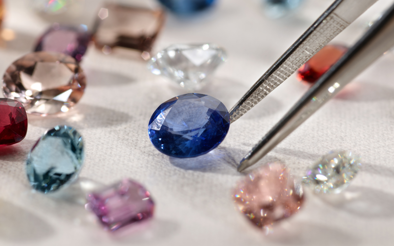 image of loose gemstones being picked by tweezers. A blue sapphire gemstone surrounded by many other colors and shapes of gemstones