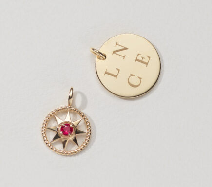 The image features two gold pendants, each with a distinct design and style, laid out against a plain, light-colored background. Left Pendant: This pendant is circular with a decorative border resembling a twisted rope. Inside the circle, there is a starburst design with an eight-pointed star. At the center of the star, there is a round red gemstone, likely a ruby, which adds a vibrant focal point to the pendant. Right Pendant: This pendant is also circular but features a smooth, polished surface. The surface is engraved with the letters "L N C E," arranged in a square pattern, giving it a personalized and meaningful touch. The pendant has a simple and elegant design, suitable for engraving initials or other significant letters. The overall presentation highlights the craftsmanship and unique designs of each pendant, showcasing their potential as meaningful and stylish accessories. The combination of classic and contemporary elements makes them versatile pieces that can be worn for various occasions.