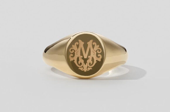 The image showcases a gold signet ring with an elegant and classic design. Material: The ring is made of polished gold, giving it a luxurious and timeless appearance. Design: The top of the ring features an engraved ornate letter "M" surrounded by intricate scrollwork, adding a personalized and sophisticated touch. The face of the ring is round and slightly raised, providing a prominent surface for the engraving. The band of the ring is smooth and tapers slightly as it wraps around the finger, ensuring a comfortable fit. The background is plain and light-colored, which helps to highlight the ring and its details. The overall presentation emphasizes the craftsmanship and elegance of the signet ring, showcasing its potential as a meaningful and stylish accessory. This piece combines classic style with personal significance, making it suitable for both everyday wear and special occasions.