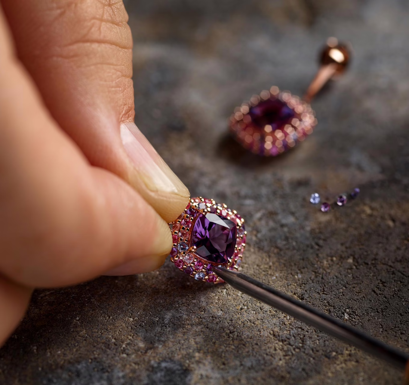 The image depicts a close-up of a person working on a piece of jewelry. The person's hand is holding a piece of jewelry set with a large purple gemstone at the center, surrounded by smaller pink and purple stones. They are using a tool, possibly a pair of tweezers or a small prying tool, to adjust or set one of the smaller stones into the piece. The background shows a textured surface, possibly a workbench, with a few loose small gemstones scattered nearby. The overall setting suggests a meticulous and detailed process of jewelry making or repair.