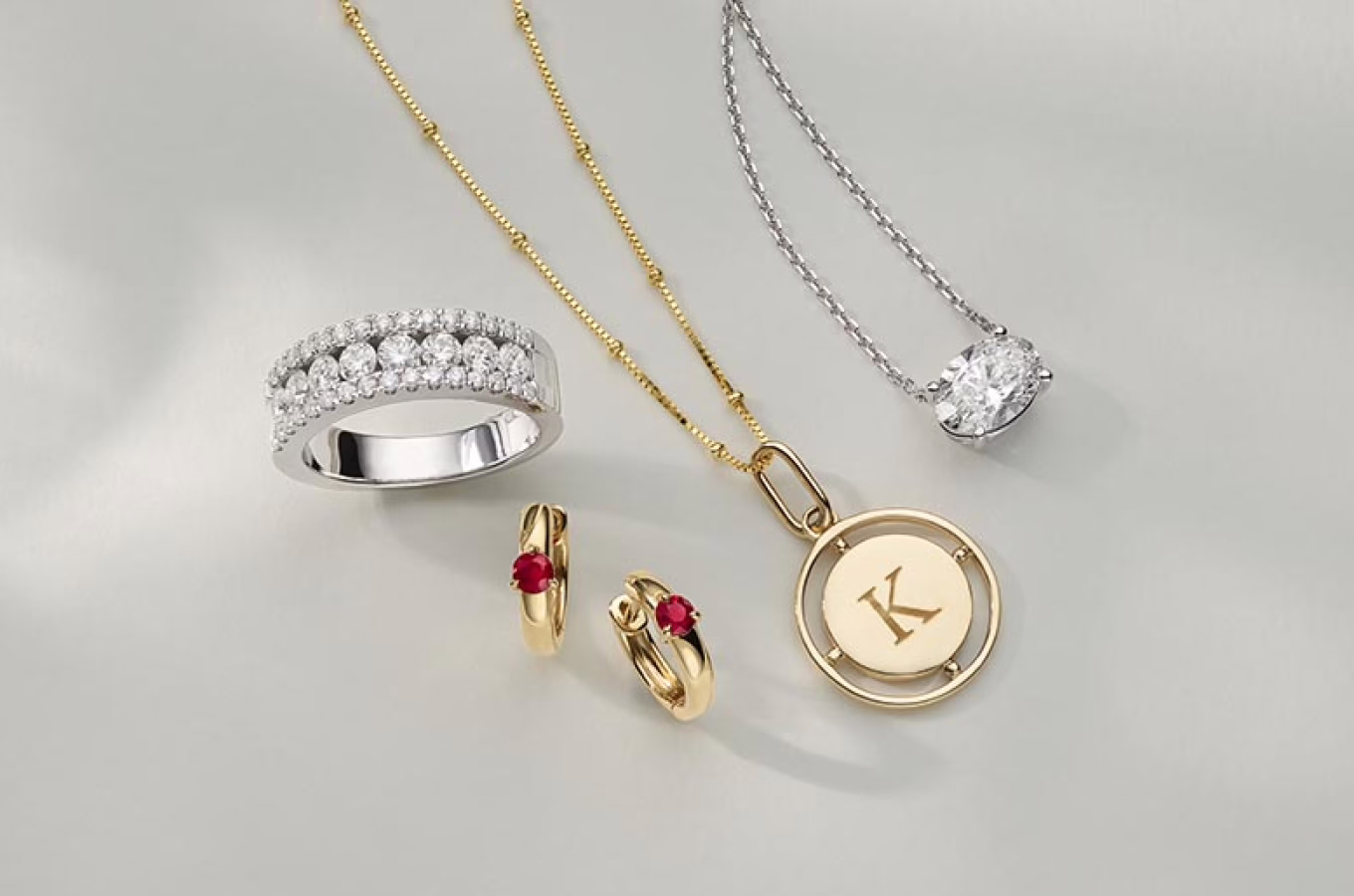 The image showcases an arrangement of various pieces of jewelry against a plain background. The collection includes: A gold ring set with multiple rows of clear, sparkling gemstones. Two necklaces: one with a gold chain and a pendant with the letter "K" engraved on it, and another with a silver chain and a large, clear gemstone pendant. A pair of gold earrings each adorned with a small red gemstone. The jewelry pieces are artfully arranged, possibly for a display or a promotional photograph, to highlight their designs and craftsmanship. The image emphasizes the elegance and luxury of the jewelry items.