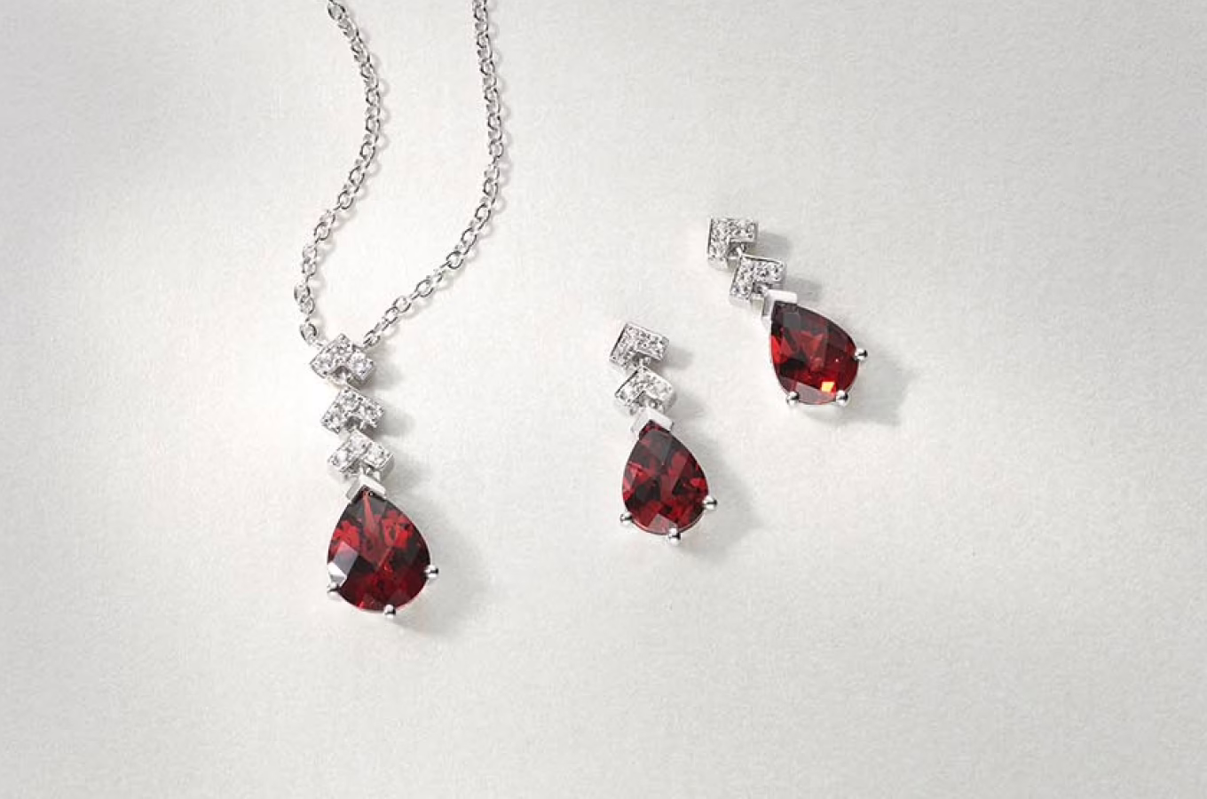 The image displays a matching jewelry set consisting of a necklace and a pair of earrings. The necklace features a silver chain with a pendant that includes a series of small clear gemstones arranged in a linear pattern, culminating in a large, teardrop-shaped red gemstone. The earrings are designed to match the necklace, each featuring the same sequence of small clear gemstones leading to a teardrop-shaped red gemstone. The background is plain and light-colored, providing a neutral backdrop that highlights the elegance and color contrast of the jewelry pieces. The overall presentation suggests a sophisticated and coordinated set, likely intended for formal or special occasions.