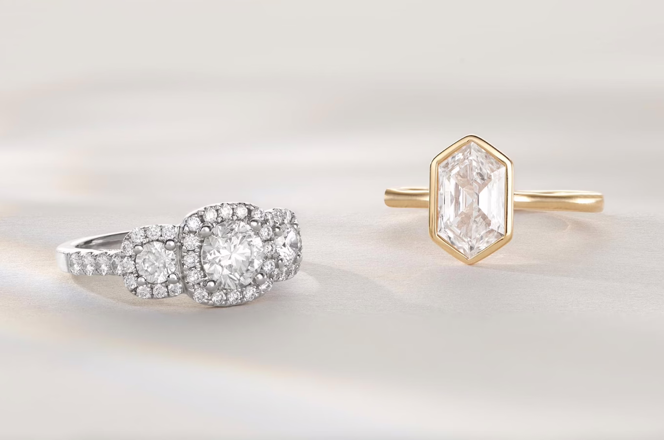 The image features two elegant rings displayed side by side against a light, neutral background. Left Ring: This ring has a white gold or platinum band and is adorned with multiple clear gemstones. At the center is a large, round diamond surrounded by a halo of smaller diamonds, enhancing its sparkle. The central diamond is flanked by two smaller round diamonds, each also surrounded by a halo of smaller diamonds. The band itself is encrusted with additional small diamonds, creating a dazzling and intricate design. Right Ring: This ring has a thin gold band and features a unique hexagonal-shaped clear gemstone, likely a diamond, at its center. The diamond is set in a bezel style, with the gold metal rim encasing its perimeter. The elongated hexagonal cut of the diamond showcases its facets and brilliance. The design is modern and sleek, with the simplicity of the band drawing attention to the distinctive shape and size of the gemstone. Together, these rings present a contrast in styles: one being more traditional and ornate with multiple diamonds, and the other being modern and minimalist with a focus on the unique shape of the central gemstone. The overall presentation highlights the craftsmanship and elegance of each piece.