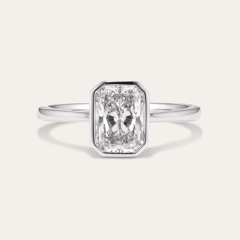 The image displays a close-up of an elegant ring. The ring features a thin, polished white gold or platinum band and is set with a prominent, rectangular-shaped clear gemstone, likely a diamond. The gemstone is cut in a radiant or emerald style, showcasing its facets and brilliance. It is secured in a bezel setting, with the metal rim encasing and protecting the edges of the diamond, giving the ring a modern and sleek appearance. The background is a plain, light beige color, which helps to highlight the ring and its details. The overall presentation is simple and sophisticated, emphasizing the clarity and sparkle of the diamond as well as the refined craftsmanship of the ring. This piece appears to be designed as a statement or engagement ring, combining timeless elegance with contemporary design elements.