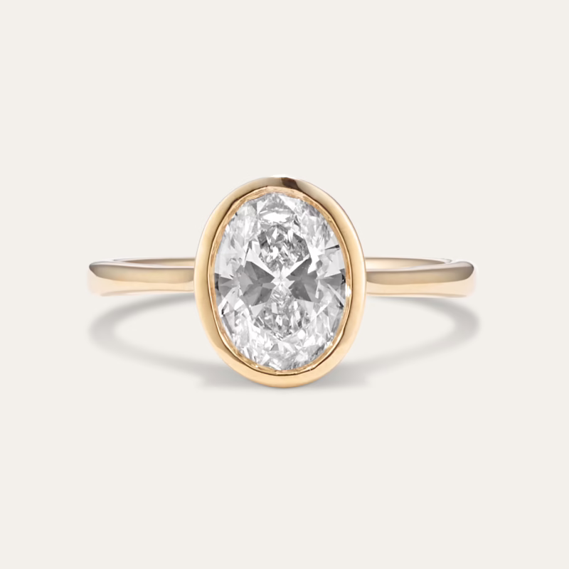 The image features a close-up view of an elegant ring. The ring has a thin gold band and is set with a prominent, oval-shaped clear gemstone, likely a diamond. The gemstone is encased in a bezel setting, meaning it is surrounded by a continuous metal rim, rather than being held by prongs. This type of setting provides a sleek and modern look, ensuring the gemstone is securely held in place. The background is a plain, light beige color, which helps to emphasize the ring and its details. The overall presentation is simple yet sophisticated, highlighting the brilliance and clarity of the diamond as well as the fine craftsmanship of the ring. This piece appears to be a statement piece or an engagement ring, designed to capture attention with its elegant and timeless design.
